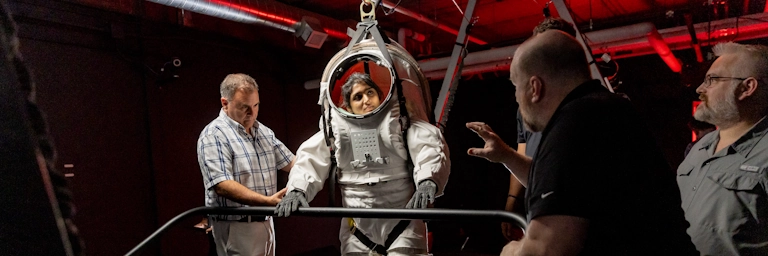 A student performs tests in a spacesuit, while faculty explain what to do